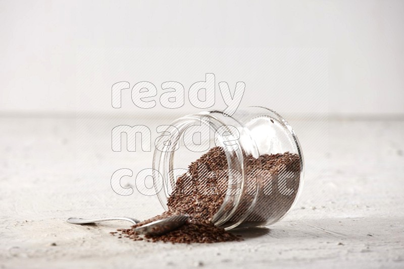 A glass jar full of flax seeds flipped and seeds spread out with a metal spoon full of the seeds on a textured white flooring