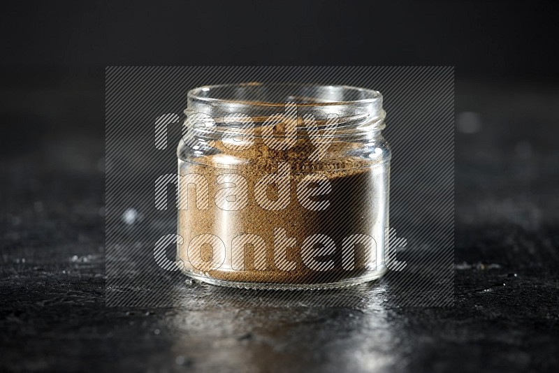 A glass jar full of cumin powder on a textured black flooring