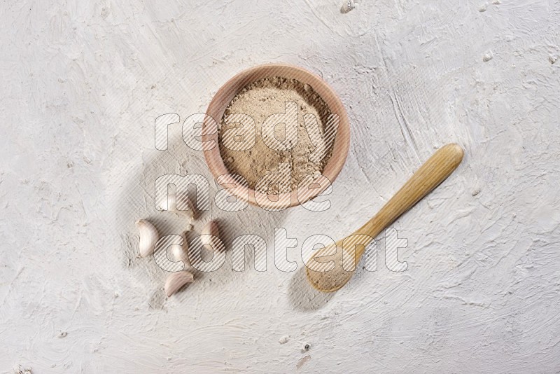 A wooden bowl and spoon full of garlic powder and beside it garlic cloves on a textured white flooring in different angles
