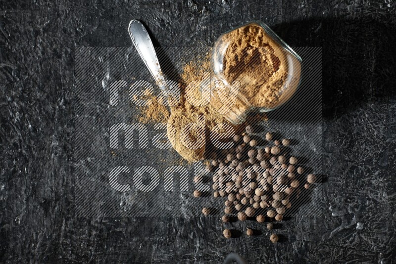 A flipped glass spice jar and metal spoon full of allspice powder and powder spilled out of it with whole balls on a textured black flooring