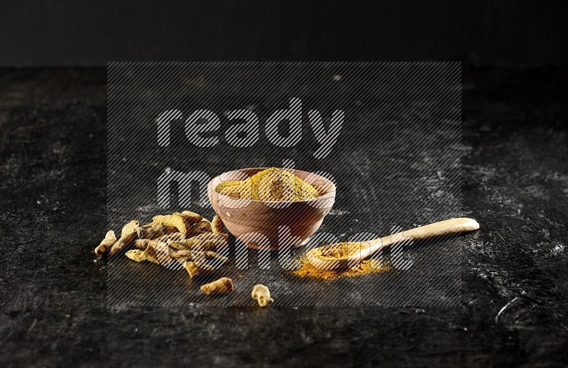 A wooden bowl and a wooden spoon full of turmeric powder with dried turmeric fingers on textured black flooring