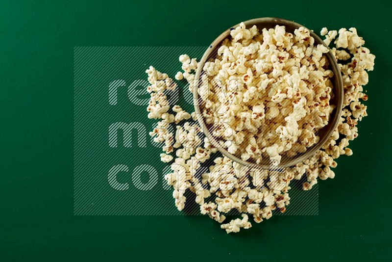 A beige ceramic bowl full of popcorn with popcorn beside it on a green background in a top view shot
