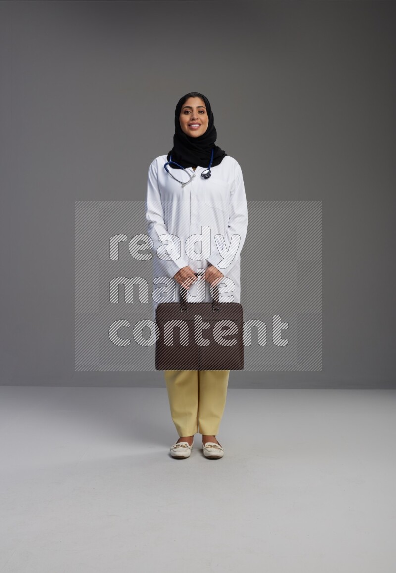 Saudi woman wearing lab coat with stethoscope standing holding bag on Gray background