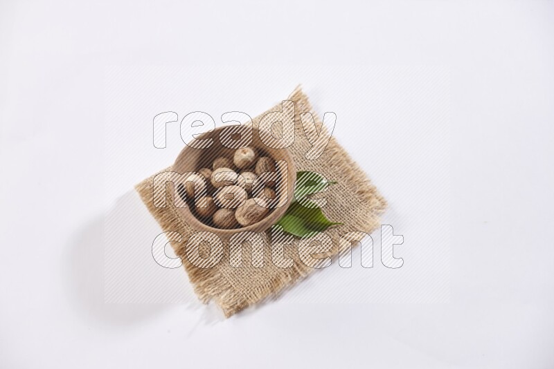 A wooden bowl full of whole nutmeg seeds on burlap fabric on a white flooring
