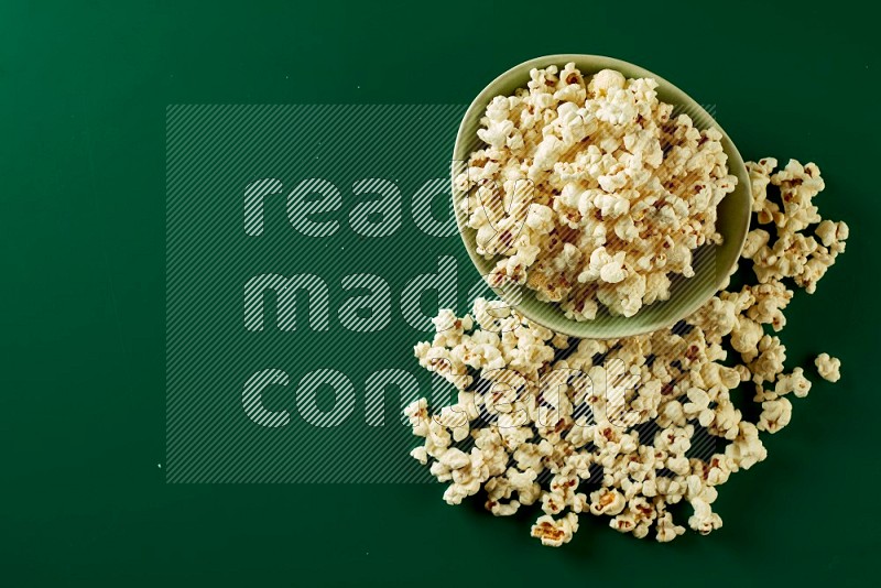 A green ceramic bowl full of popcorn with popcorn beside it on a green background in a top view shot