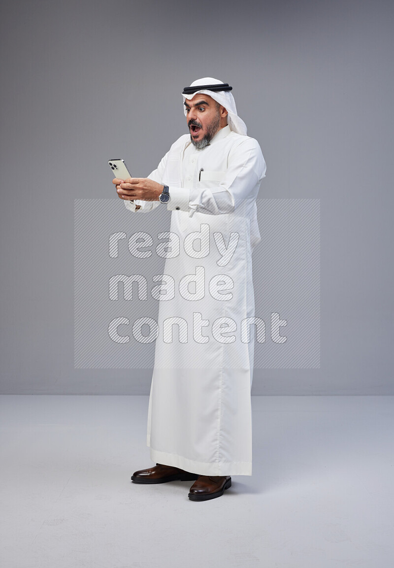 Saudi man Wearing Thob and white Shomag standing texting on phone on Gray background