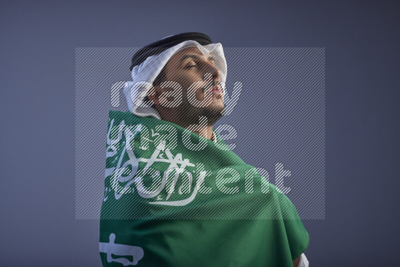 A close-up shot of Saudi man wearing thob and white shomag wrapping big Saudi flag on gray background