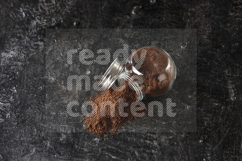 A flipped glass spice jar full of cloves powder and powder came out of it on textured black flooring