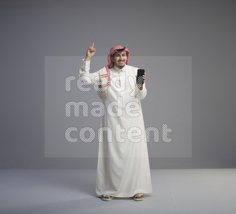 A Saudi man standing wearing thob and red shomag showing phone to camera on gray background
