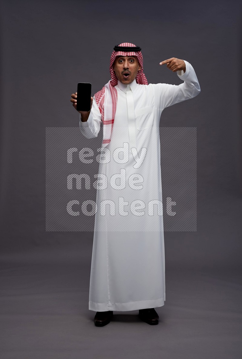 Saudi man wearing thob and shomag standing showing phone to camera on gray background