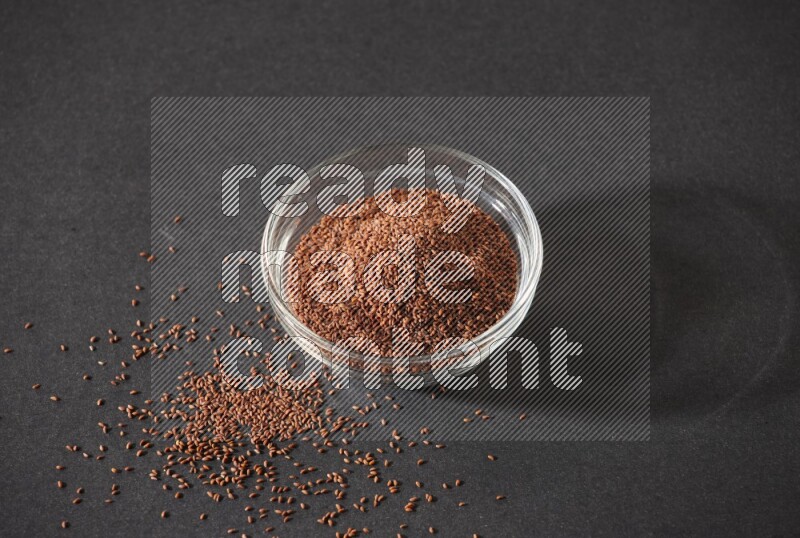 A glass bowl full of garden cress seeds surrounded by seeds on a black flooring