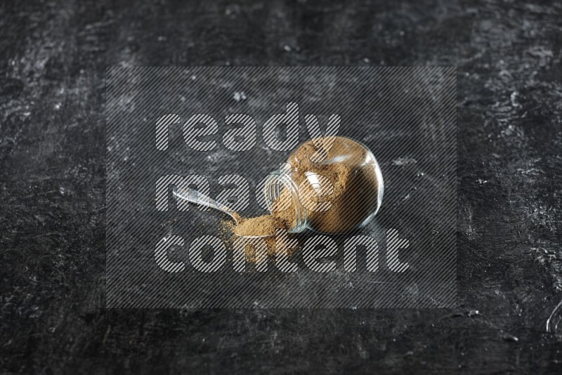 وعاء زجاجي للتوابل مقلوب وملعقة معدنية ممتلئة ببودرة الكمون والبودرة تناثرت منه على خلفية سوداء