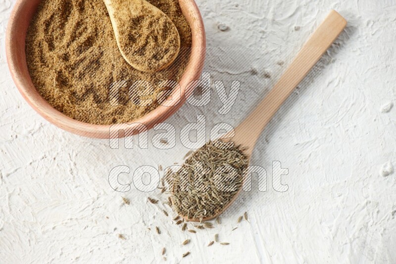 A wooden bowl and 2 wooden spoons full of cumin powder and cumin seeds on textured white flooring