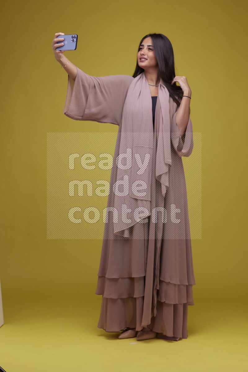A woman Taking a Selfie on a Yellow Background wearing Brown Abaya