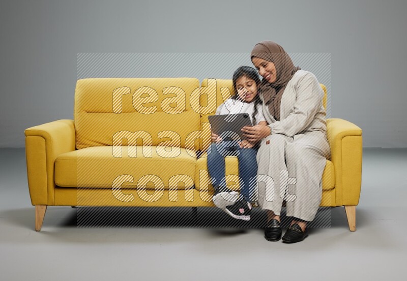 A girl with her mother on yellow sofa and watching on iPad on gray background