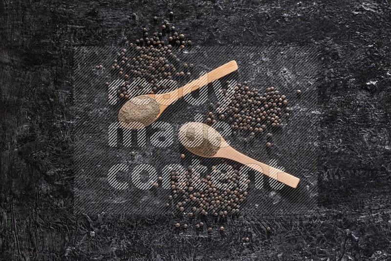 2 wooden spoons full of black pepper powder and black pepper beads spread on a textured black flooring