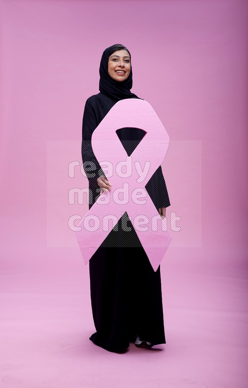 Saudi woman wearing pink ribbon on abaya standing holding awareness ribbon on plain pink background