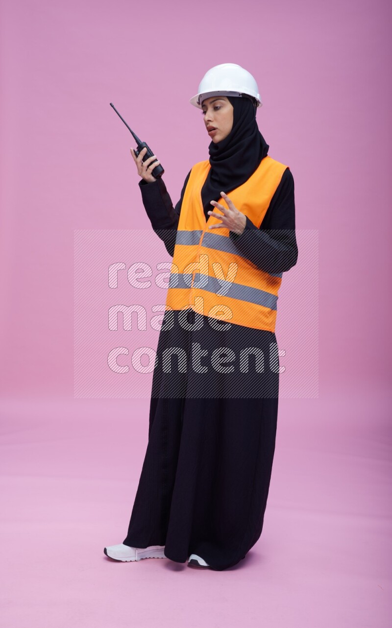 Saudi woman wearing Abaya with engineer vest and helmet standing holding walkie-talkie on pink background