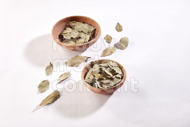 2 wooden bowls full of dried bay leaves with more leaves spread on white flooring