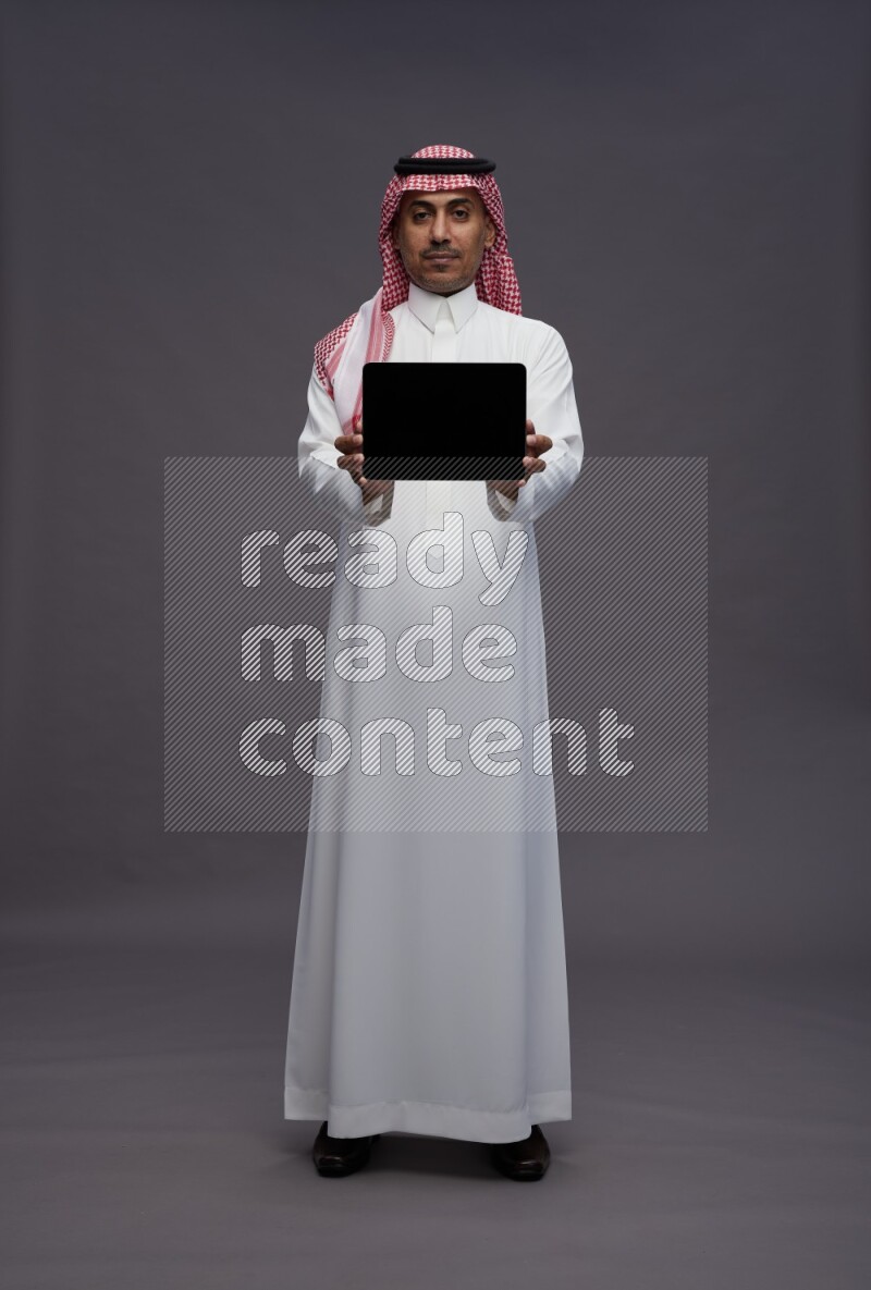 Saudi man wearing thob and shomag standing showing tablet to camera on gray background