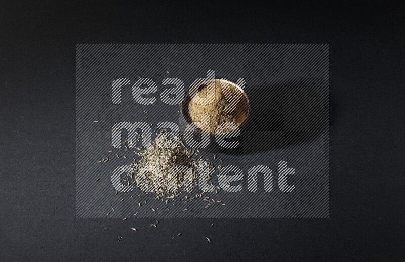 وعاء فخاري أسود ممتلئ ببودرة الكمون وحبوب الكمون متناثرة على خلفية سوداء