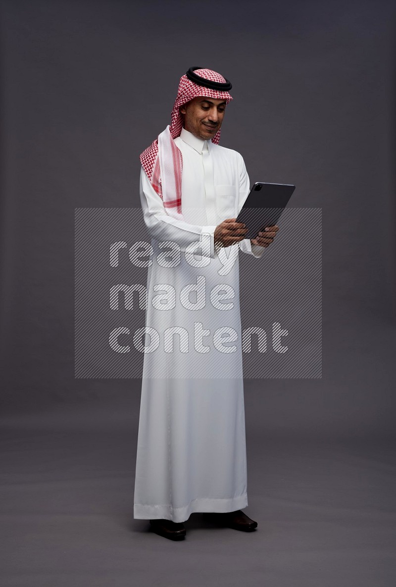 Saudi man wearing thob and shomag standing working on tablet on gray background