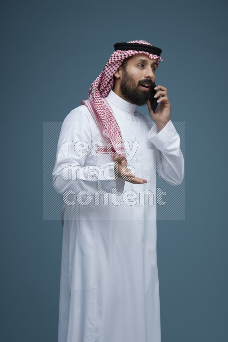 A man having a Call in a blue background wearing Saudi Thob with Shomag