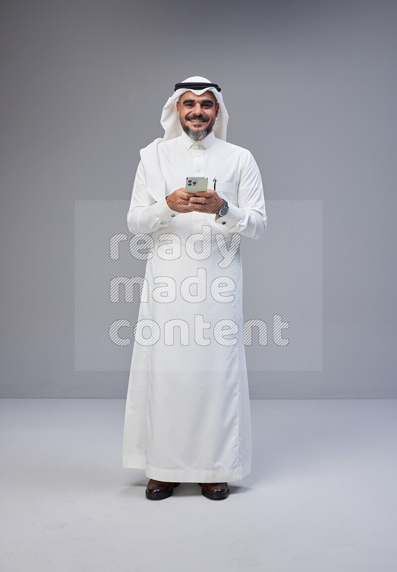 Saudi man Wearing Thob and white Shomag standing texting on phone on Gray background