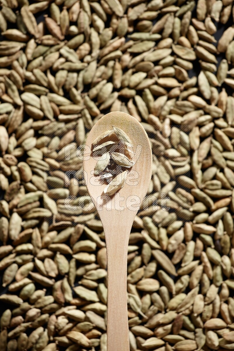 Cardamom seeds in a wooden spoon above cardamom seeds background on black flooring