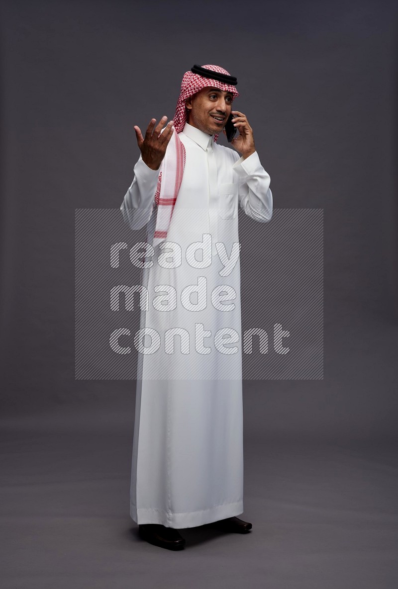 Saudi man wearing thob and shomag standing talking on phone on gray background