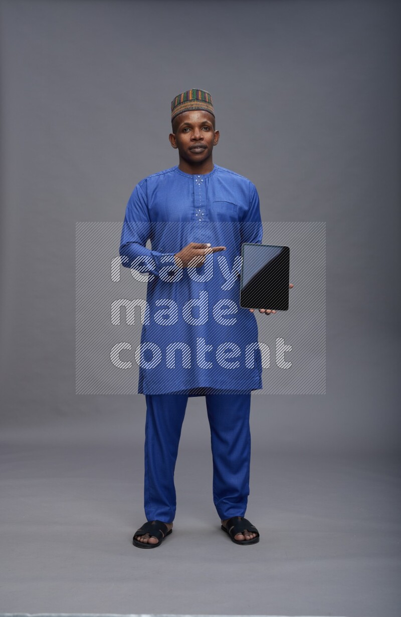 Man wearing Nigerian outfit standing showing tablet to camera on gray background