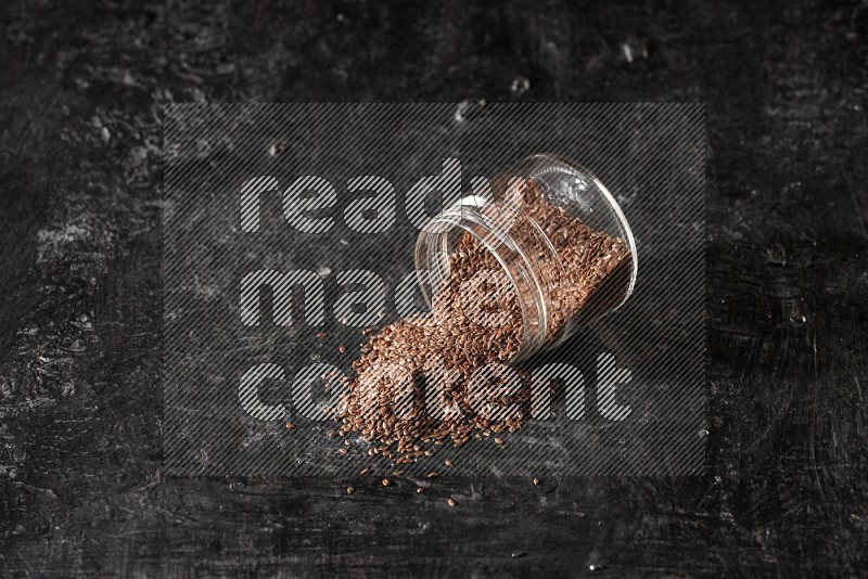 A glass jar full of flaxseeds flipped and seeds spread out on a textured black flooring