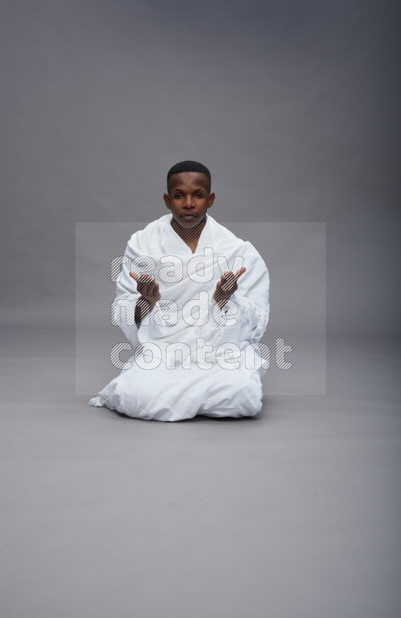 A man wearing Ehram sitting on floor dua'a on gray background