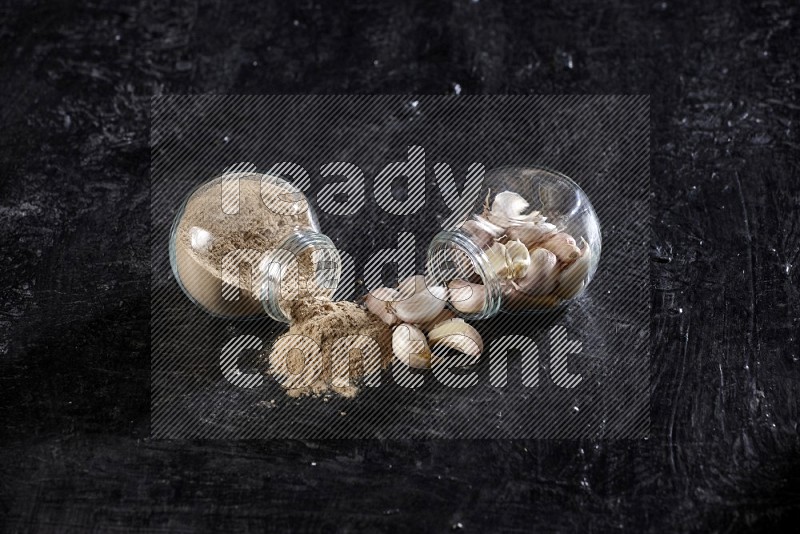 2 glass spice jars, one full of garlic powder and the other full of garlic cloves both flipped on a textured black flooring