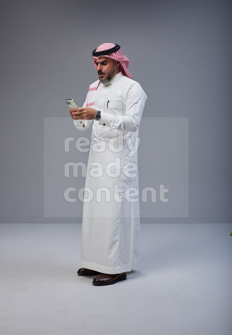 Saudi man Wearing Thob and red Shomag standing texting on phone on Gray background