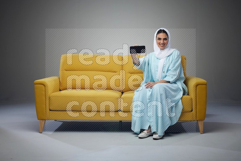 A Saudi woman wearing a light blue Abaya and white head scarf sitting on a yellow sofa and showing her phone's screen vertically and horizontally eye level on a grey background