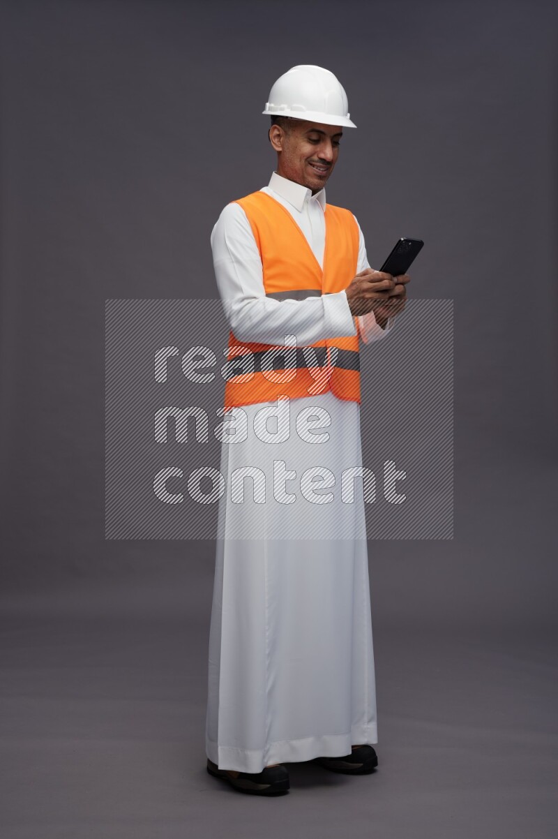 Saudi man wearing thob with engineer vest standing texting on phone on gray background