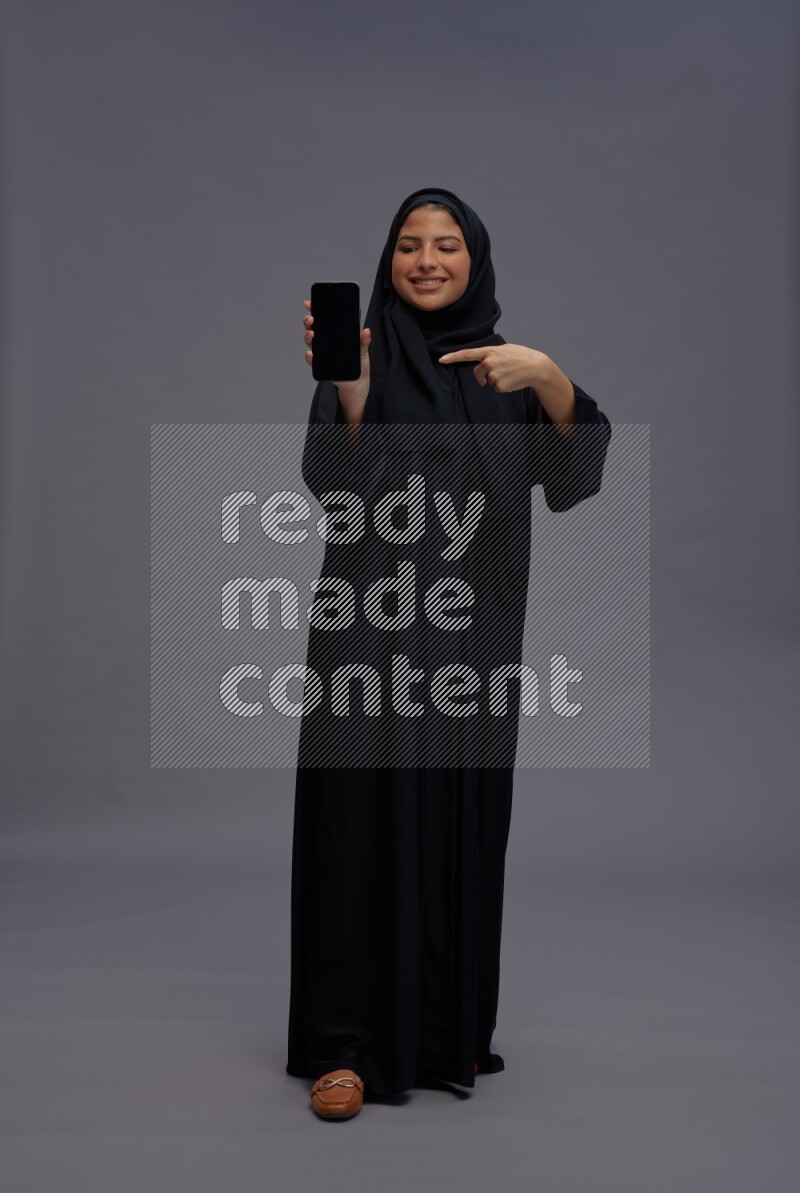 Saudi woman wearing Abaya standing showing phone to camera on gray background