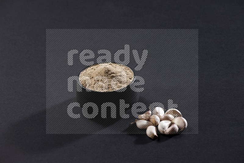 A black pottery bowl full of garlic powder and cloves beside it on a black flooring in different angles
