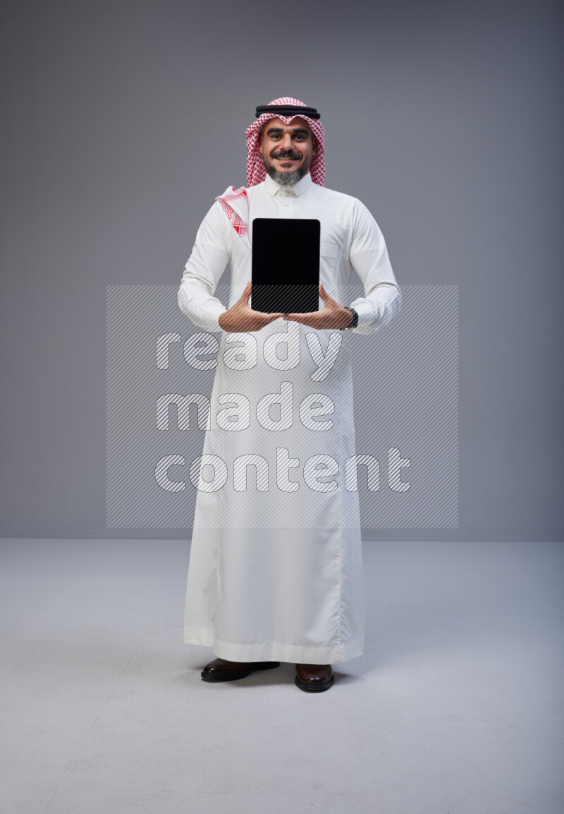 Saudi man Wearing Thob and red Shomag standing showing tablet to camera on Gray background