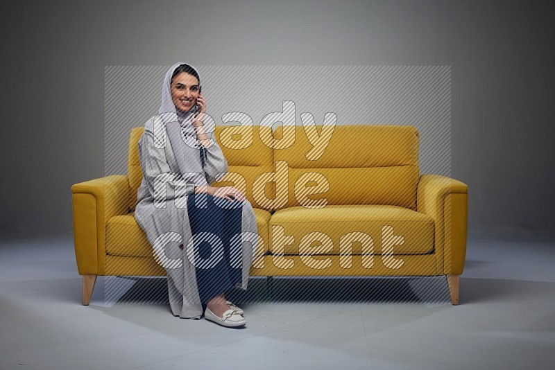 A Saudi woman wearing a light gray Abaya and head scarf sitting on a yellow sofa and talking in the phone eye level on a grey background