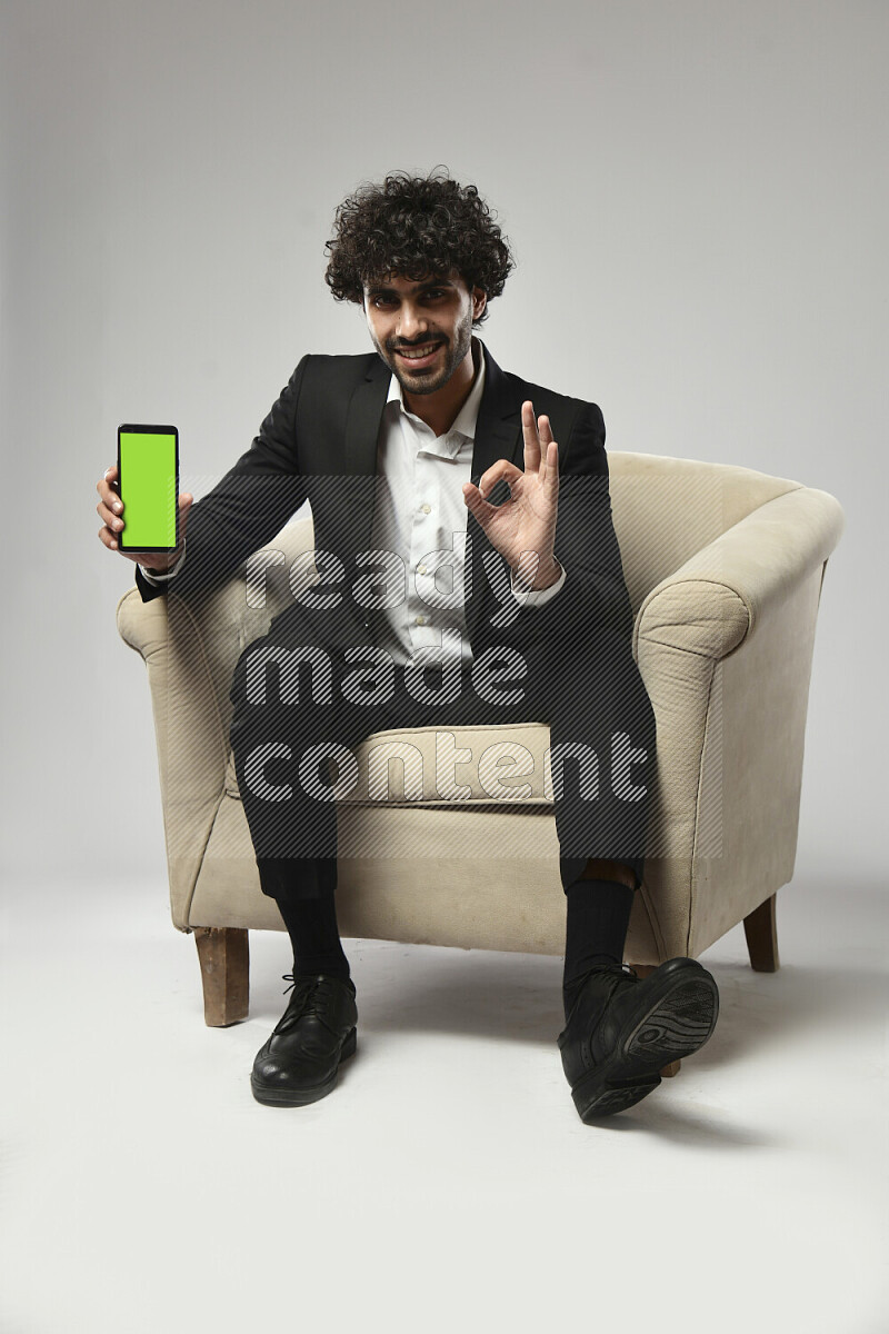 A man wearing formal sitting on a chair showing a phone screen on white background