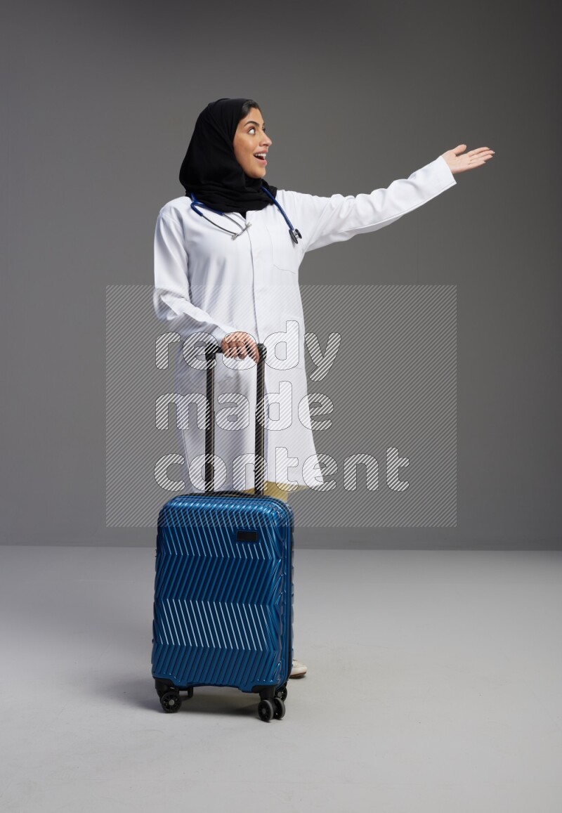Saudi woman wearing lab coat with stethoscope standing holding Travel bag on Gray background
