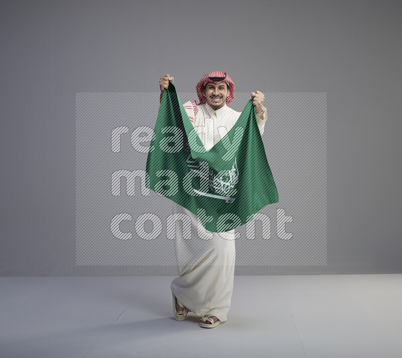 A saudi man standing wearing thob and red shomag holding big saudi flag on gray background