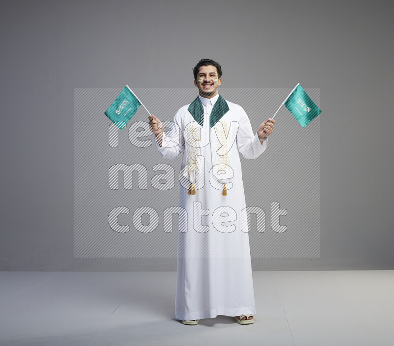 A Saudi man standing wearing thob and saudi flag scarf with face painting holding small Saudi flag on gray background