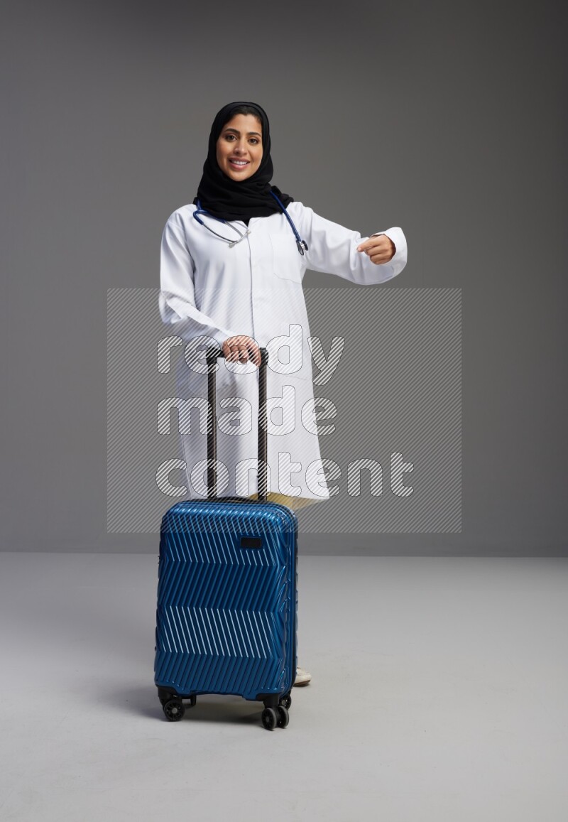 Saudi woman wearing lab coat with stethoscope standing holding Travel bag on Gray background
