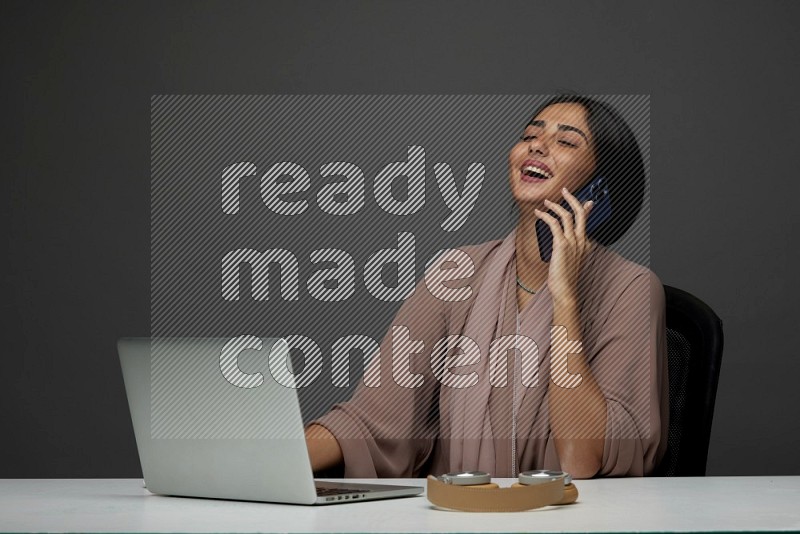 A Saudi woman Sitting on her desk Calling  on a Gray Background wearing Brown Abaya