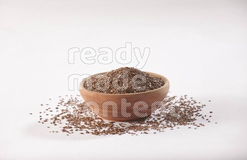 A wooden bowl full of flax seeds surrounded by flax seeds on a white flooring