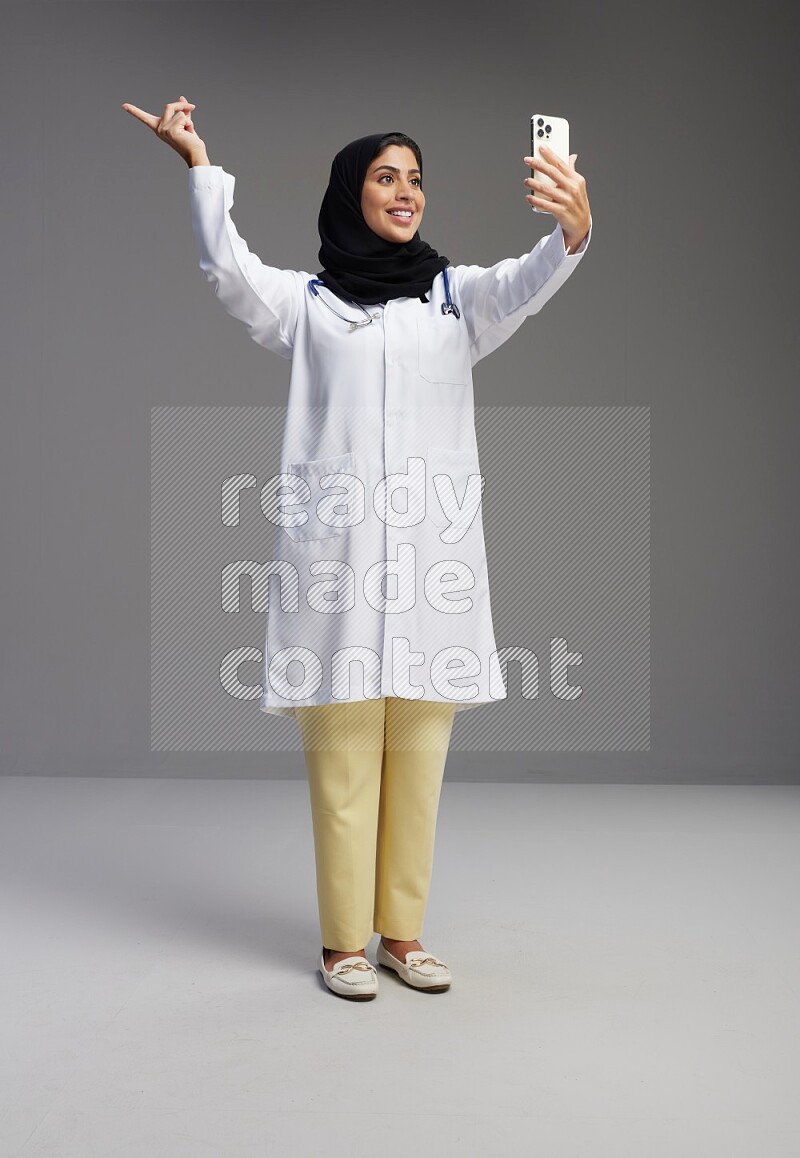 Saudi woman wearing lab coat with stethoscope standing taking selfie on Gray background