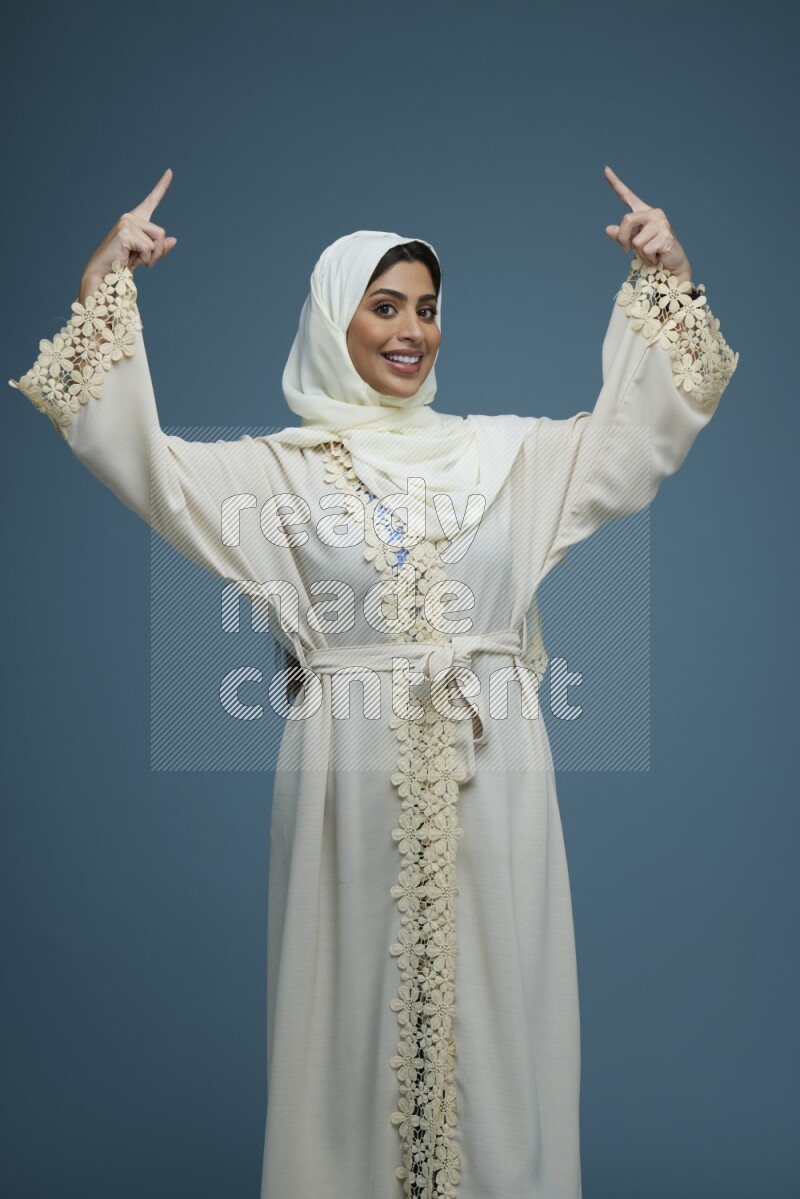 A Saudi woman pointing in a blue background wearing an off-white Abaya Hijab